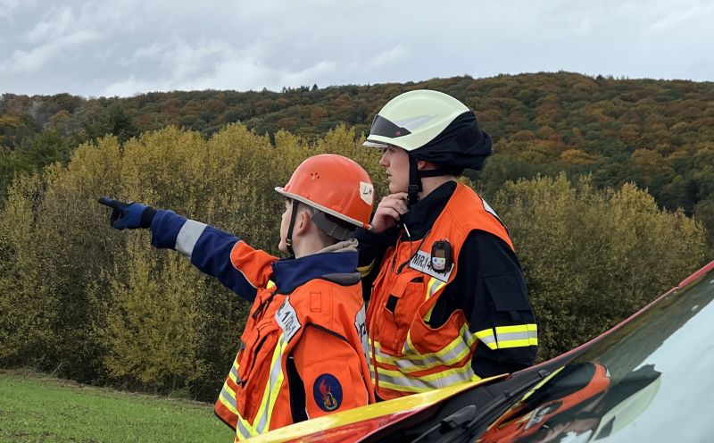 Zwei Mitglieder der Jugendfeuerwehr in orangefarbenen Schutzjacken und Helmen stehen im Freien, einer zeigt mit ausgestrecktem Arm in die Ferne, im Hintergrund herbstlich gefärbte Bäume und bewaldete Hügel.