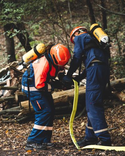 Zwei Jugendliche in Feuerwehrschutzkleidung mit Atemschutzgeräten auf dem Rücken kuppeln einen gelben Feuerwehrschlauch im Wald.