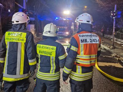 Drei Feuerwehrleute in Schutzkleidung und Helmen stehen mit dem Rücken zur Kamera vor einem Einsatzfahrzeug mit eingeschaltetem Blaulicht auf einer nächtlichen Straße