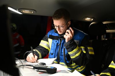 Feuerwehrmann in Schutzkleidung sitzt in einem Fahrzeug, spricht in ein Funkgerät und macht sich Notizen auf einem Klemmbrett